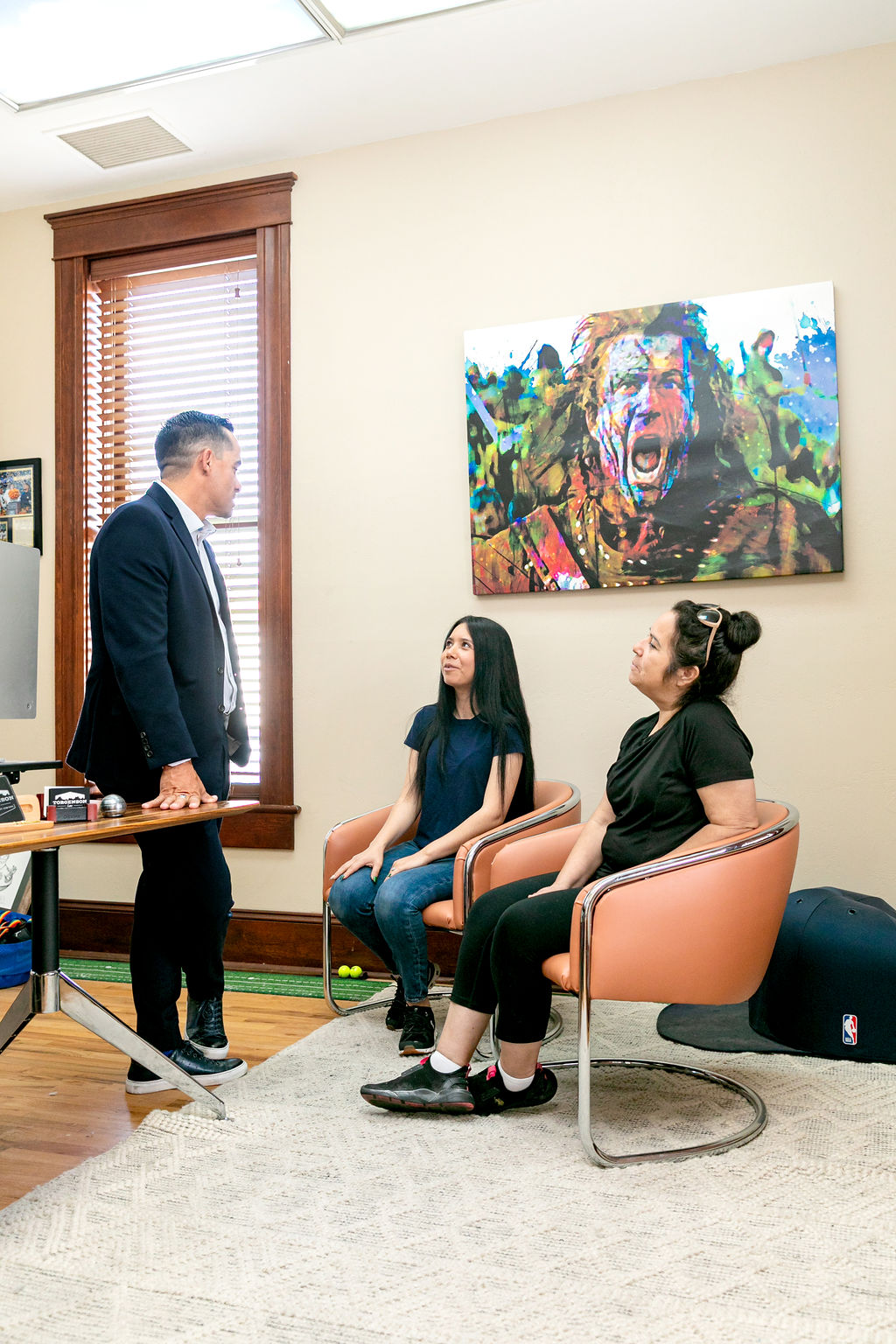 A man in a suit stands and talks to two women sitting in chairs in an office with a large, colorful painting of a shouting man on the wall.