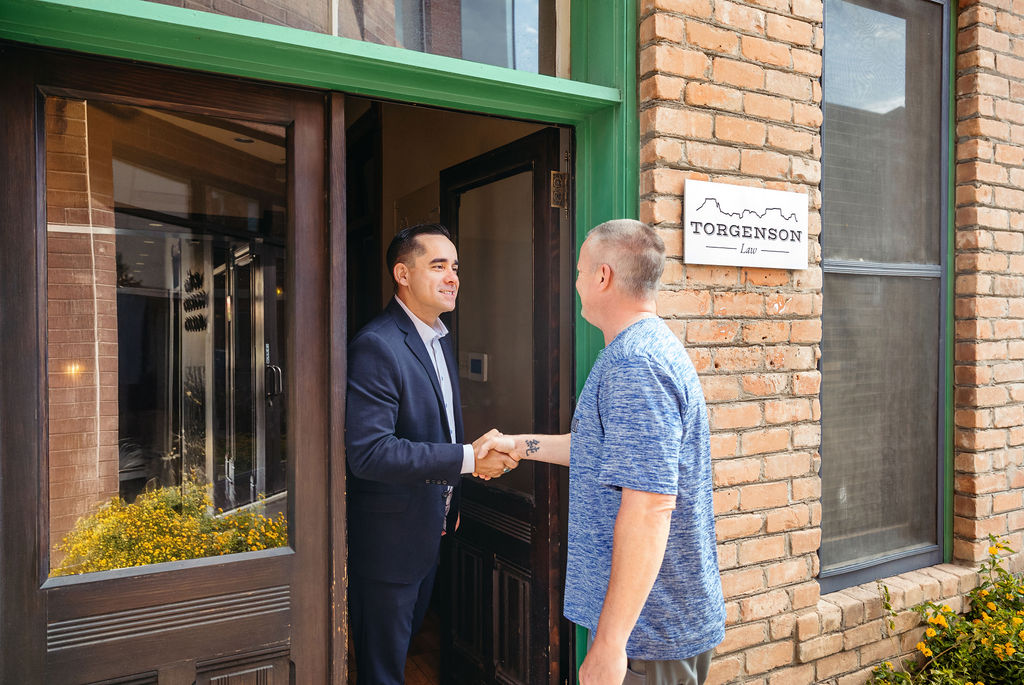 A man in a suit shakes hands with a man in a blue t-shirt in the doorway of Torgenson Law.