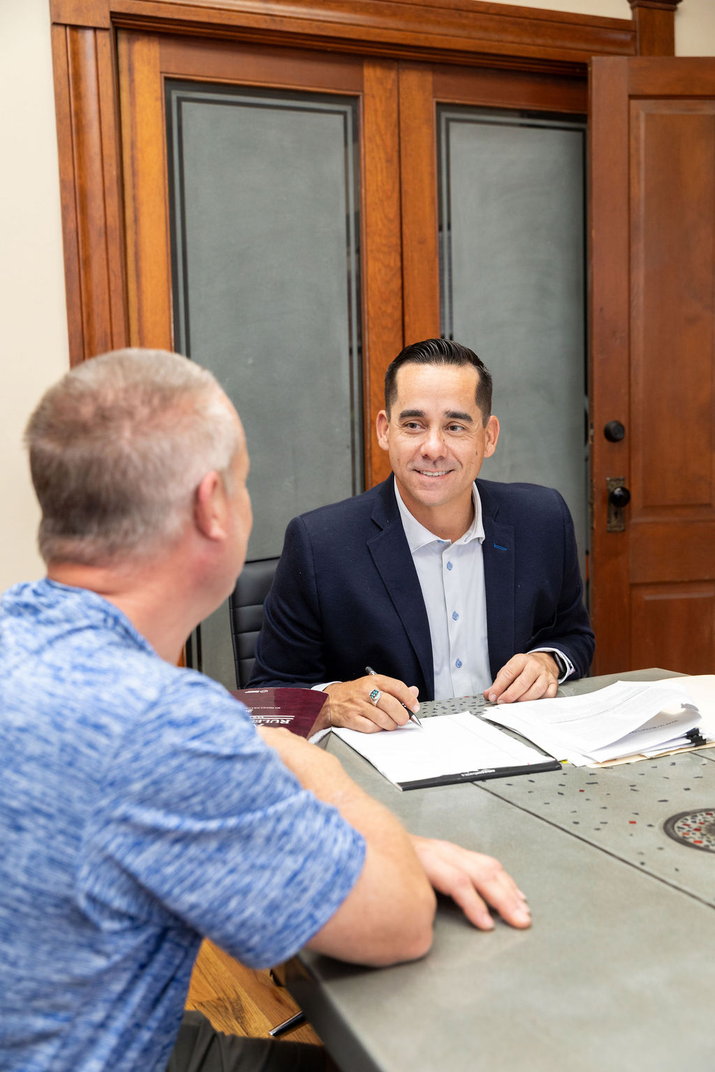 A man in a suit jacket signs a document at a table while facing a client.