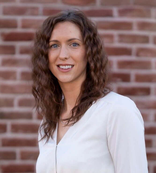 A woman with shoulder-length wavy hair smiles in front of a brick wall.