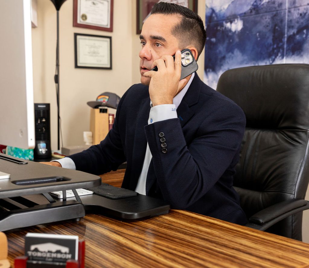 A man in a suit jacket sits at a desk while talking on a smartphone.