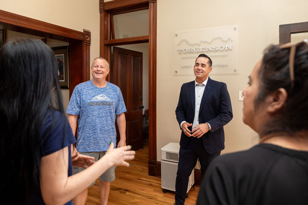 A man in a suit and a man in a t-shirt talk with two women in an office with a Torgenson sign on the wall.