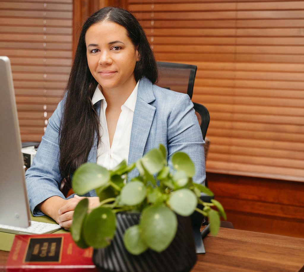 A woman in a blue blazer sits at an office desk with a potted plant and law book in the foreground.