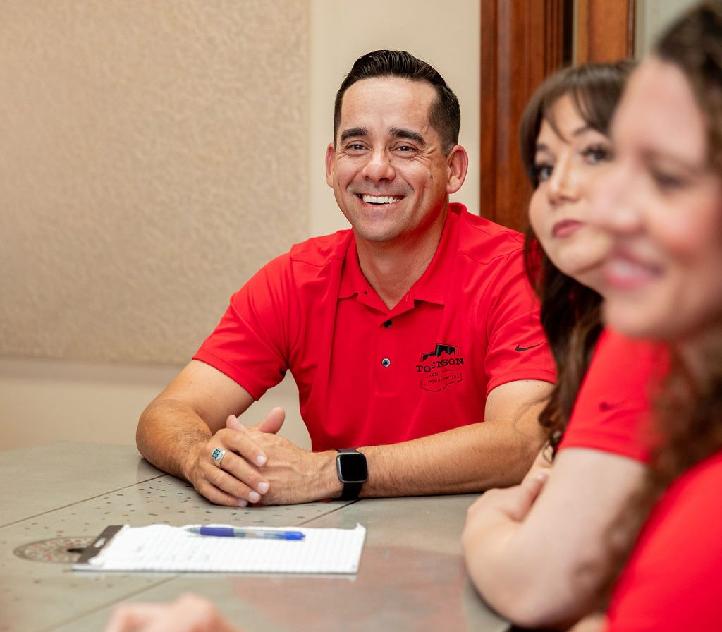 A smiling man wearing a red polo shirt sits at a conference table with two women, also in red shirts, partially visible in the foreground.