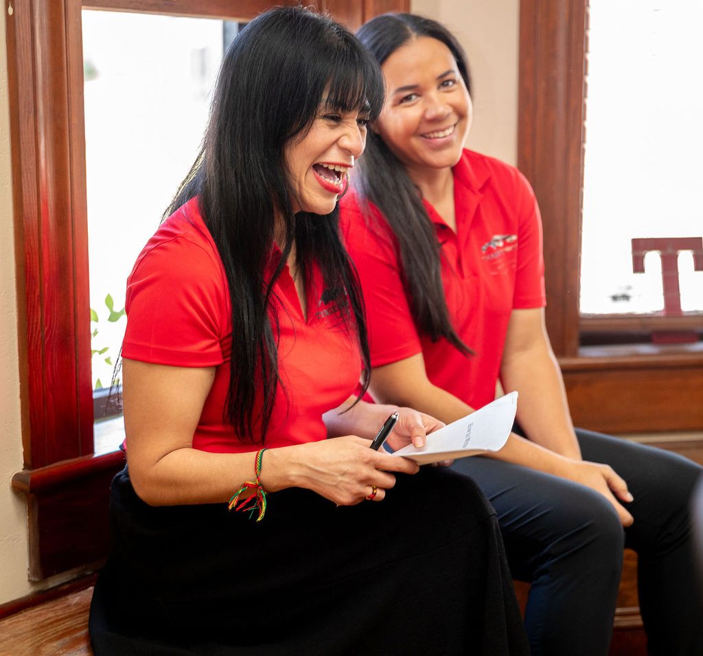 Two women in red polo shirts sit together and smile, with the woman in the foreground laughing while holding a pen and a piece of paper.