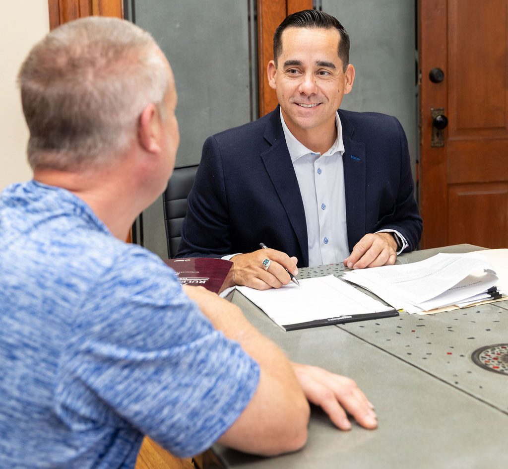 A man in a suit and dress shirt sits at a table writing on a notepad while talking to a man in a blue shirt.
