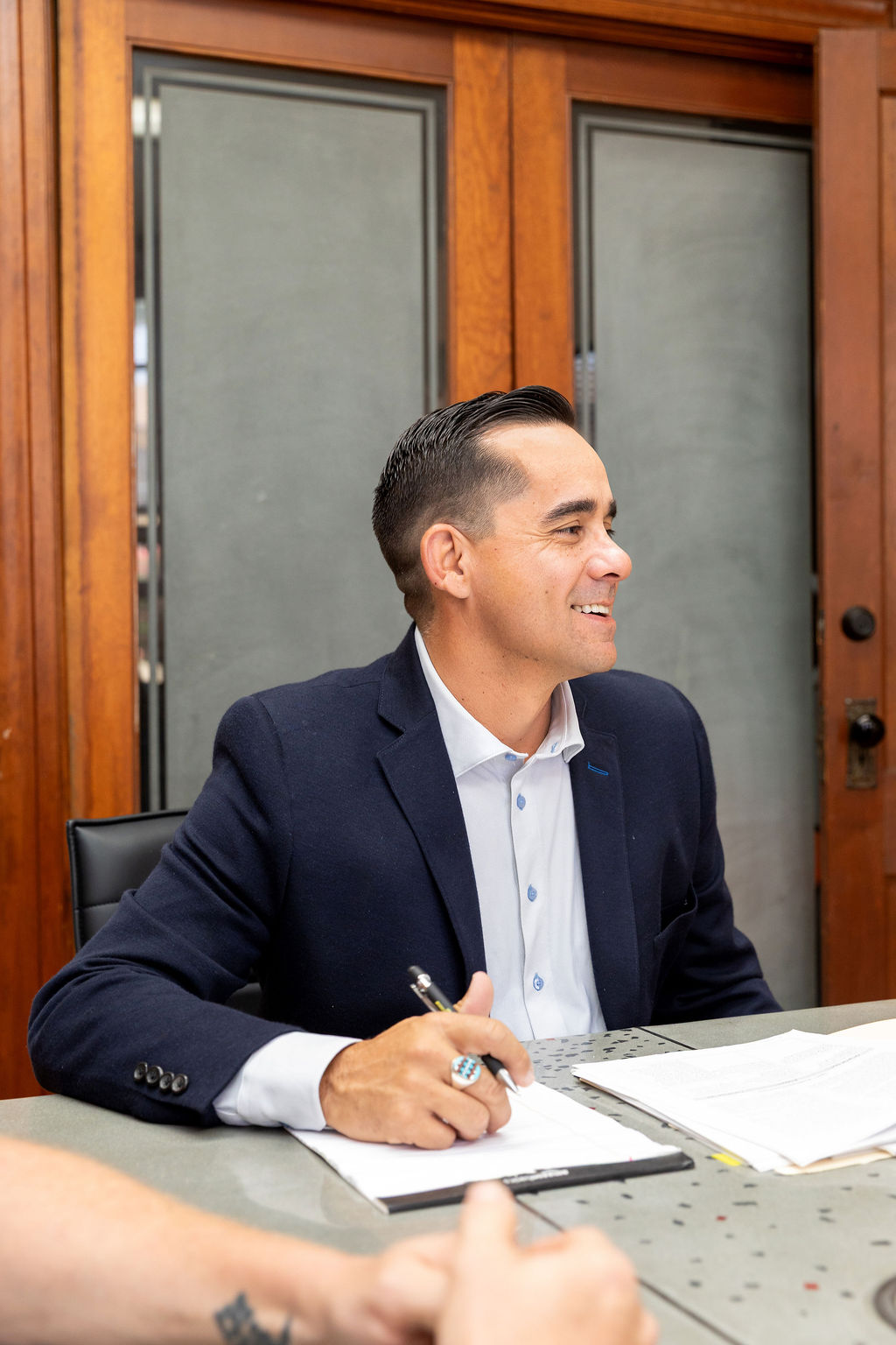 A man in a navy blazer and light shirt sitting at a table with a pen, looking to the side and smiling.