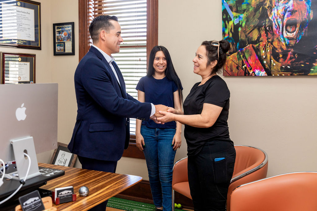 A man in a suit shakes hands with a woman in an office, as another woman smiles in the background.
