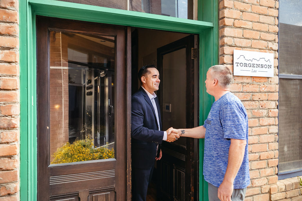 A lawyer in a suit shakes hands with a client in a blue t-shirt at the entrance of Torgenson Law.