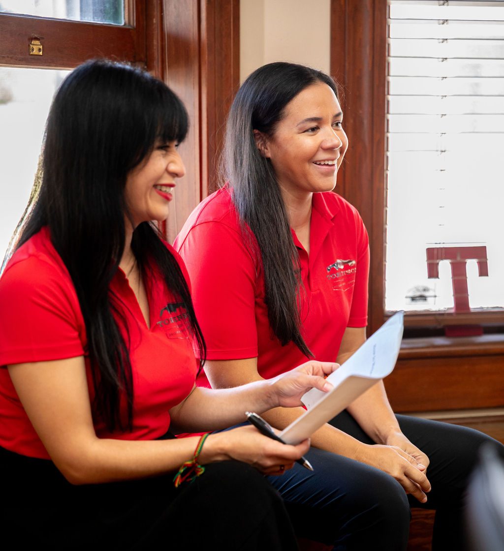 Two women in red polo shirts sit together, with one holding a document and pen while they talk.