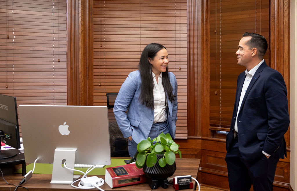 A smiling woman in a blue suit and a man in a navy suit stand in an office having a conversation.