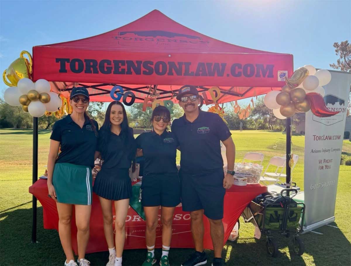 Four people wearing black shirts stand in front of a red Torgenson Law event tent on a golf course.