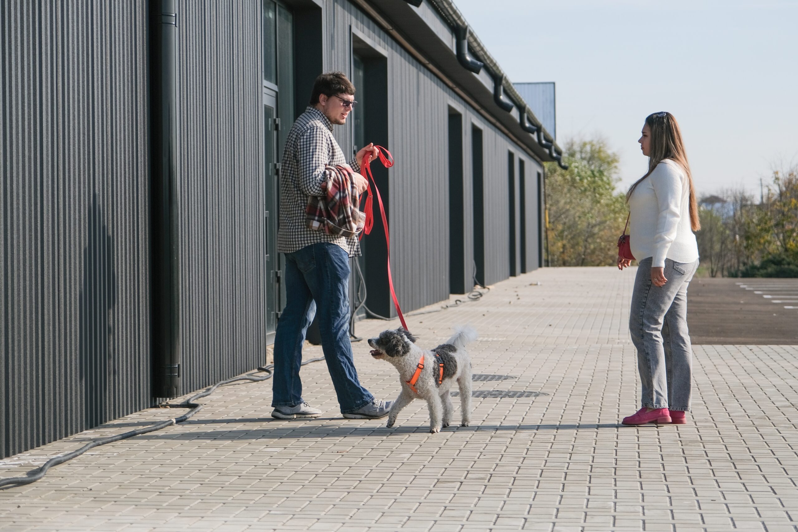A man standing with a dog on a leash and a woman standing nearby on a paved walkway outside a building looking confused