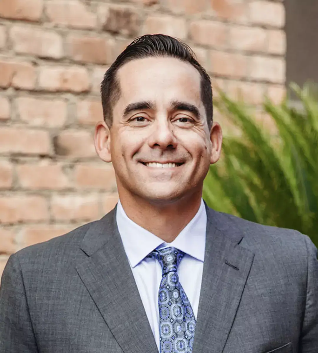A smiling man in a suit and patterned tie poses in front of a brick wall.
