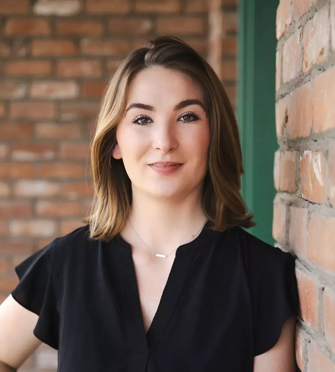 A woman with shoulder-length brown hair wearing a black blouse stands against a brick wall.