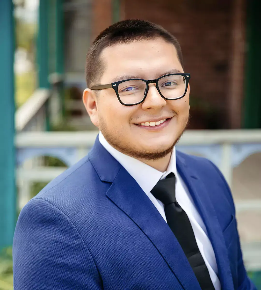 A man wearing a blue suit, white shirt, black tie, and glasses smiles at the camera.