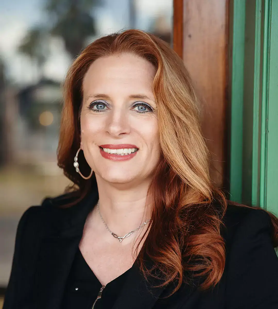 A woman with shoulder-length reddish-brown hair wearing a black jacket and hoop earrings, smiling at the camera.