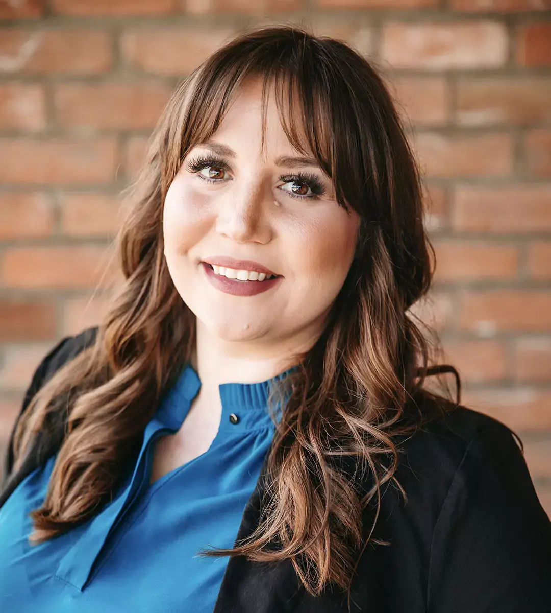 Portrait of a smiling woman wearing a blue top and a black blazer in front of a brick wall.