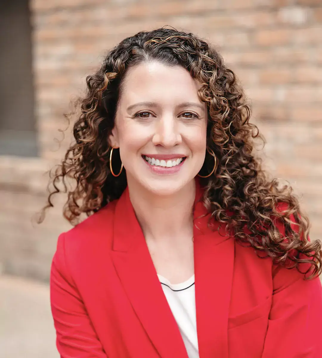 A woman with curly hair smiles while wearing a red blazer and gold hoop earrings.