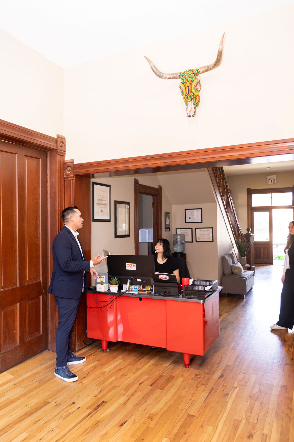 A man in a suit speaks to a woman sitting at a red desk in an office lobby, with a decorative longhorn skull mounted on the wall above them.