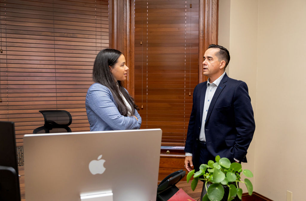 A woman and a man in business attire stand in an office talking to each other.