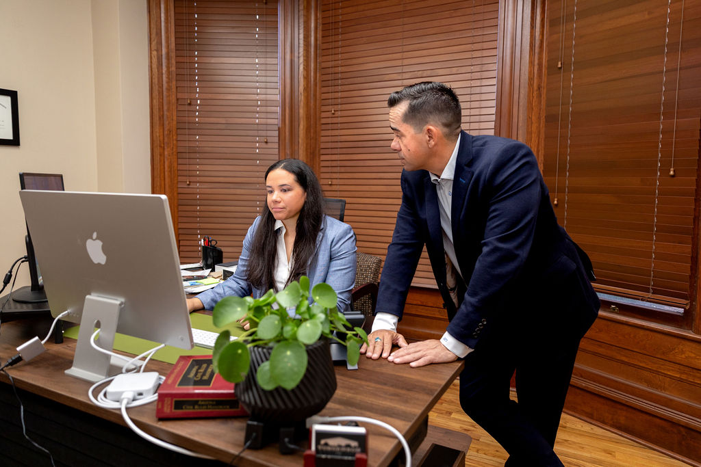 A woman sits at a desk looking at a computer monitor while a man in a suit leans over to consult with her.