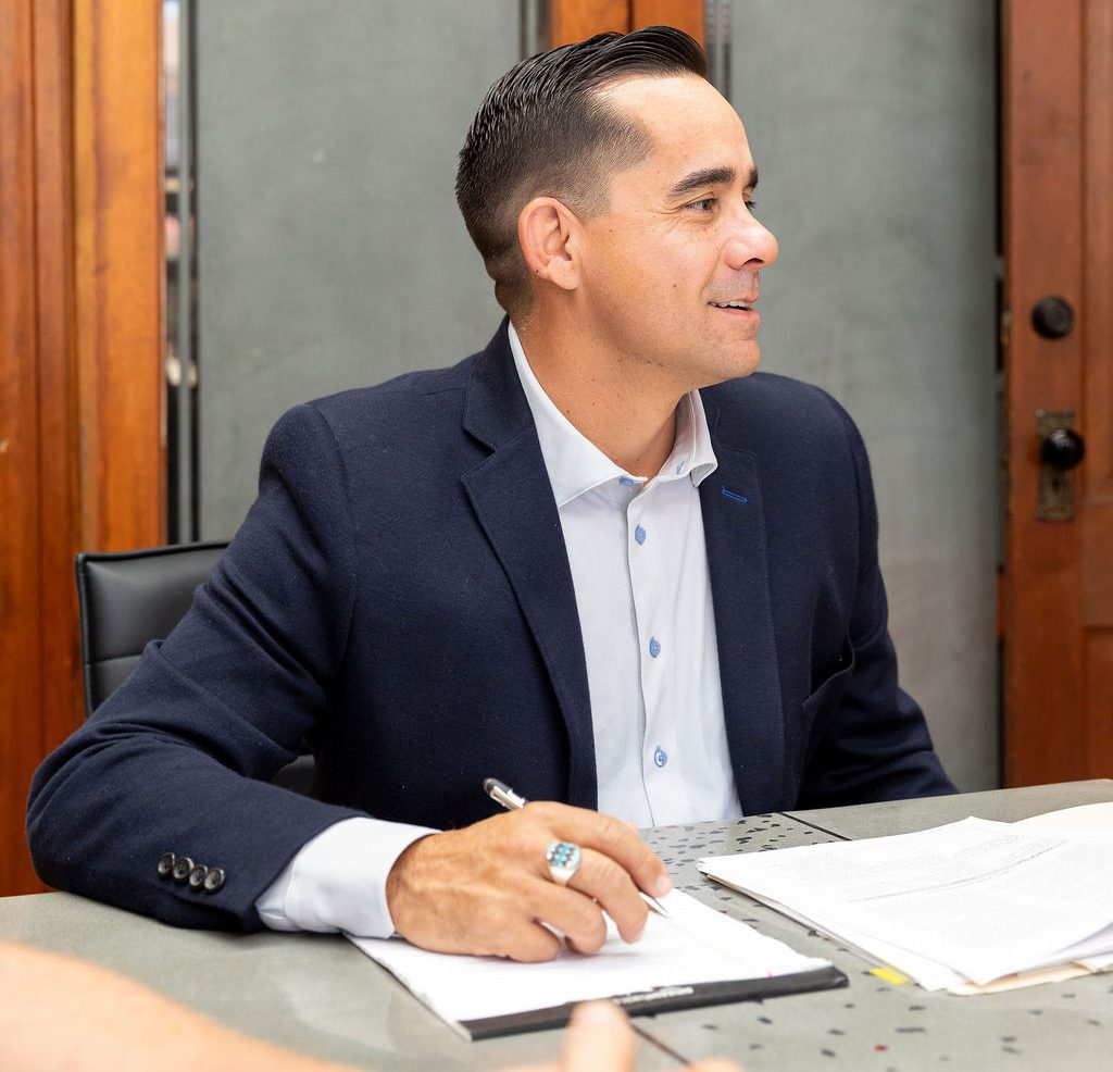 A man in a navy blazer and light shirt sitting at a table and writing on a notepad.