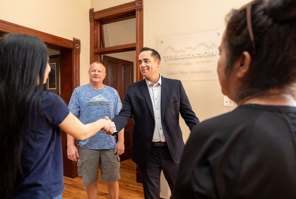 A man in a suit shakes hands with a woman while another man in a t-shirt looks on in an office with a Torgenson Law sign.