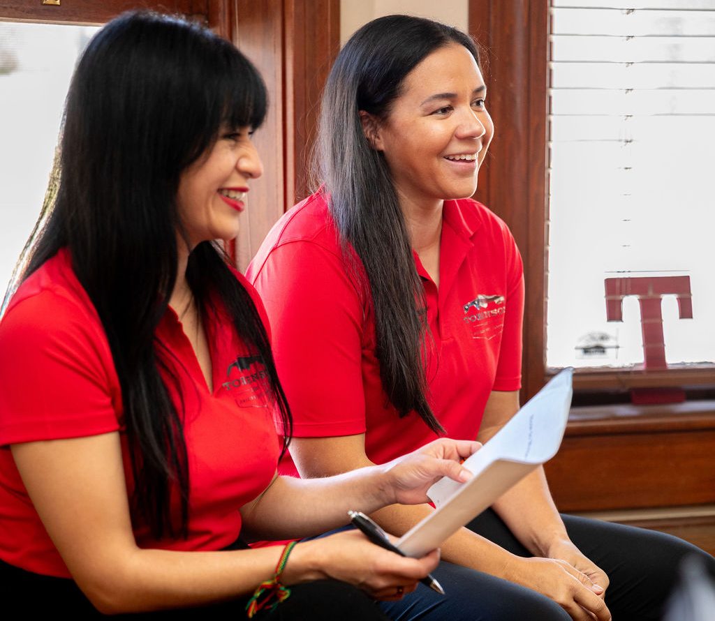 Two women in red polo shirts sit together, one holding a paper and a pen while smiling.