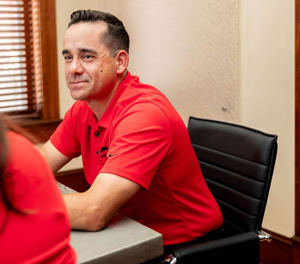 A man in a red polo shirt sits at a conference table, looking off to the side.