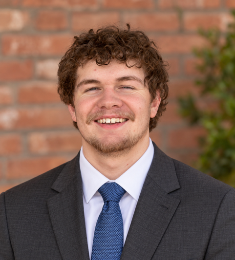 A young man with curly brown hair wearing a dark suit, white shirt, and blue tie smiles at the camera against a brick wall background.