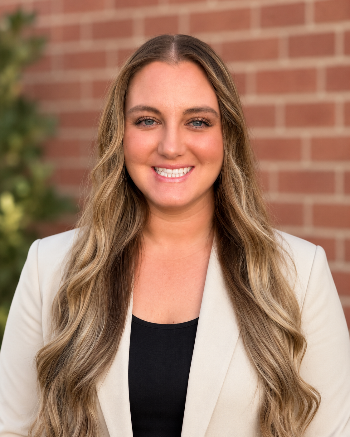 A woman with long hair wearing a white blazer over a black top smiles in front of a brick wall.